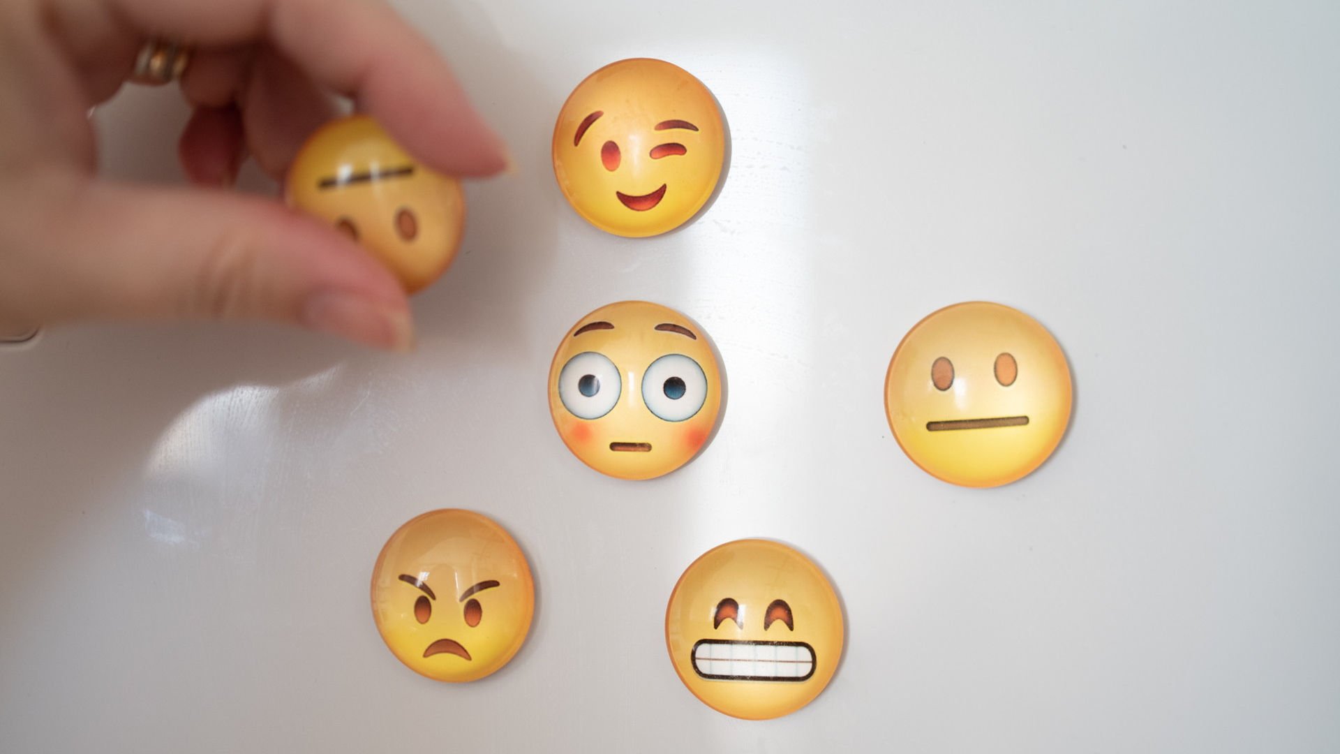 Emojis med olika uttryck på vit yta. Hand håller neutral emoji lite framför ytan.