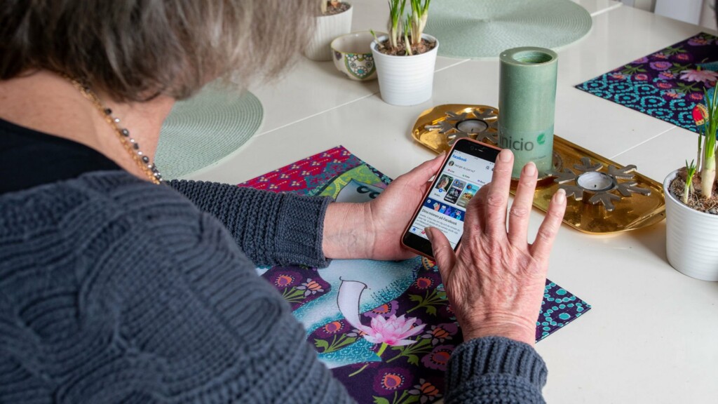En äldre kvinna sitter vid köksbordet med mobilen i handen, på skärmen syns sociala medier.