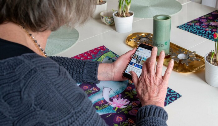 En äldre kvinna sitter vid köksbordet med mobilen i handen, på skärmen syns sociala medier.