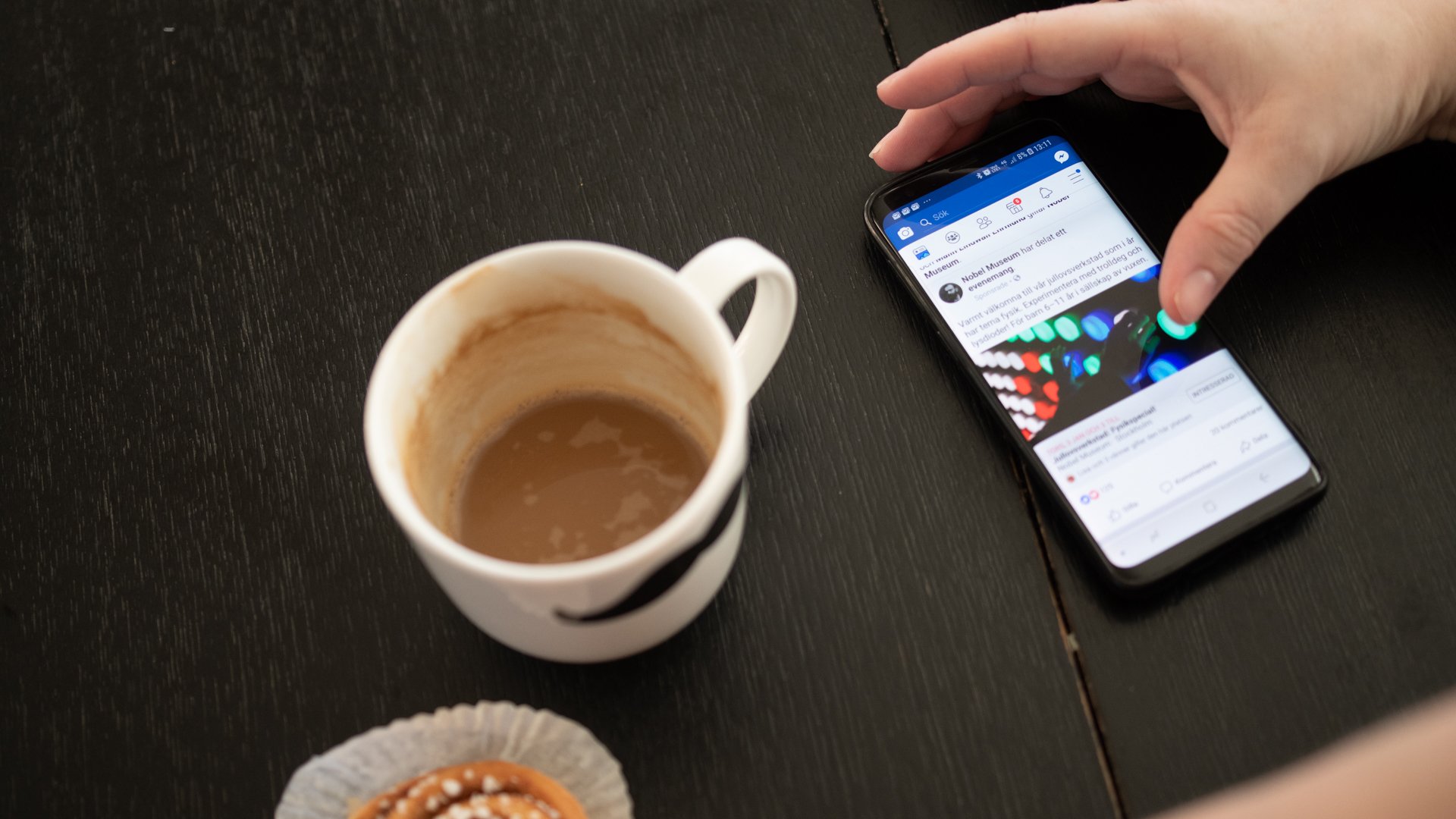 Hand använder mobiltelefon som ligger på köksbord där det även finns bulle och kaffekopp.