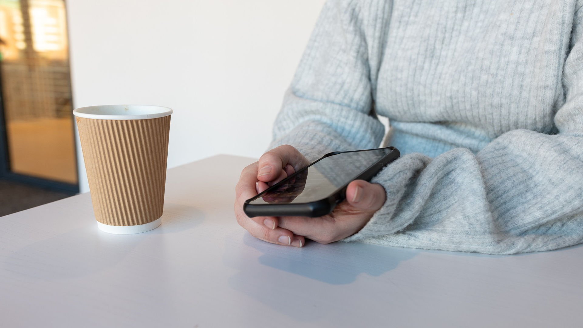 Kvinnliga händer som håller en mobiltelefon vid ett bord, bredvid står en pappersmugg.