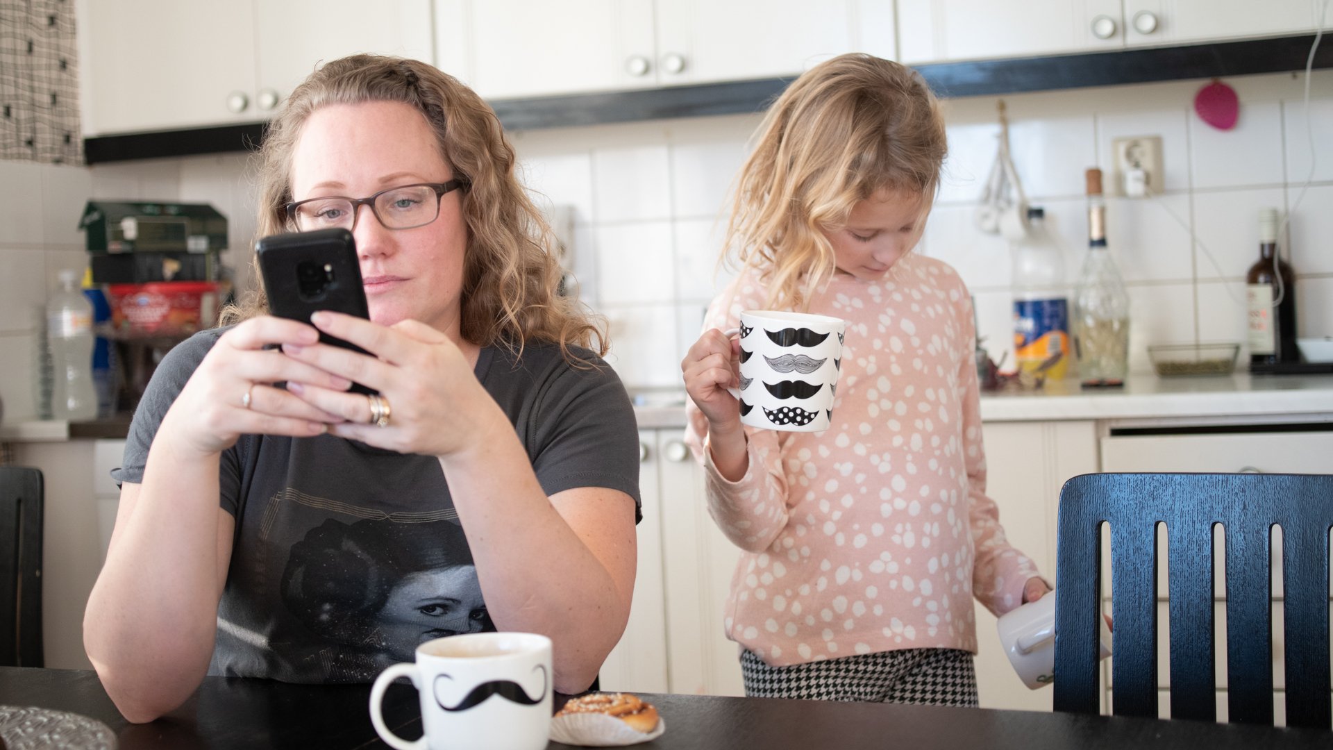 En kvinna sitter vid köksbordet och tittar på sin mobil, ett barn står bakom henne.