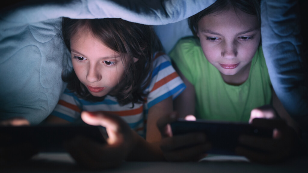 Två barn sitter under täcke och spelar på sina mobiler