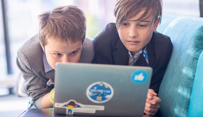 Två barn ligger på mage bredvid varandra på en soffa framför en laptop.