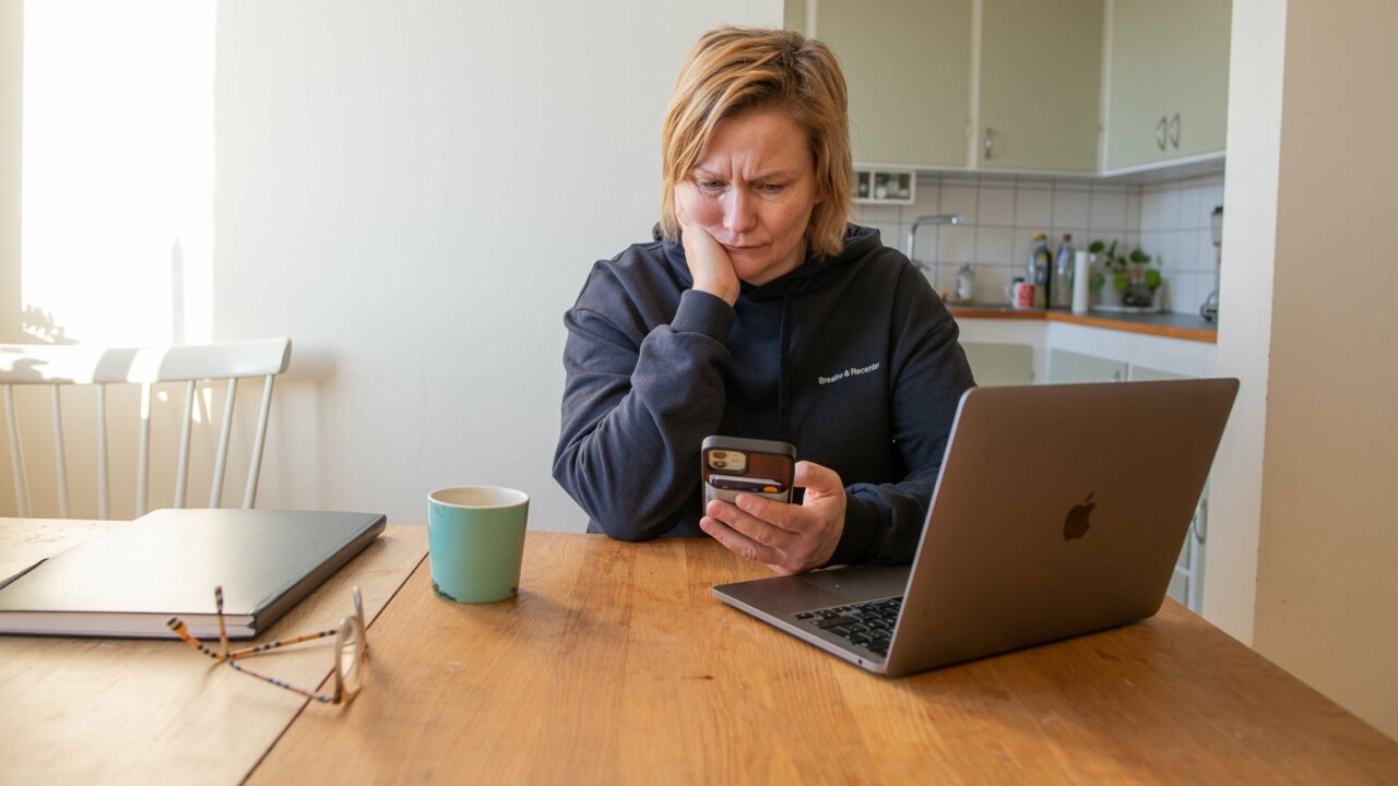 Kvinna sitter vid bord med sin mobil i handen och ser bekymrad ut