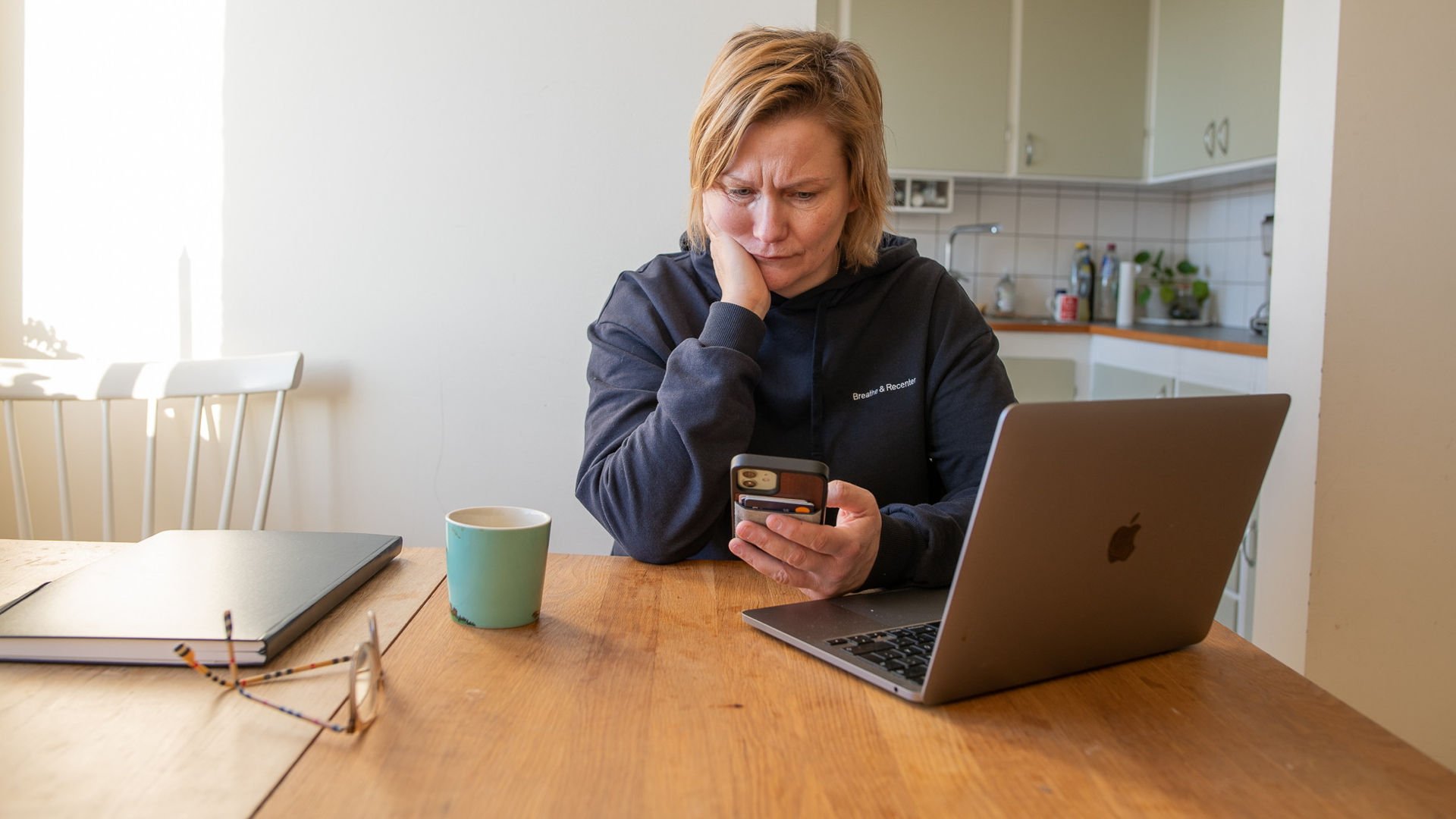 Kvinna sitter vid bord med sin mobil i handen och ser bekymrad ut