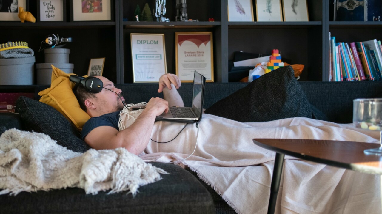 Man med hörlurar ligger under en filt i en soffa med laptop på magen.