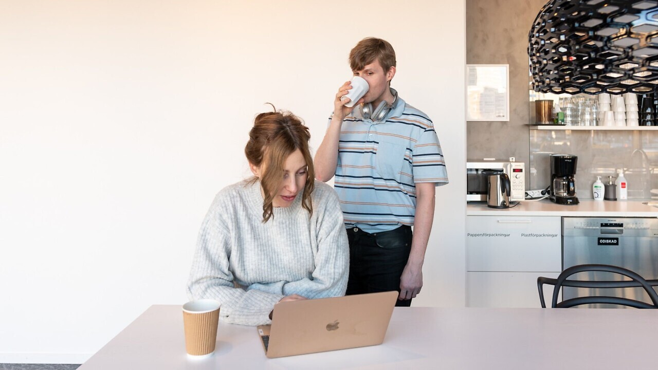 En kvinna sitter vid en laptop, en man står bakom henne och sneglar på skärmen