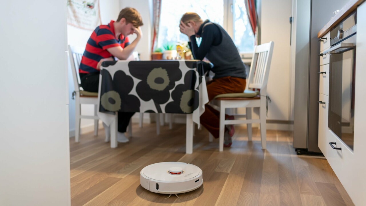 Två personer sitter vid ett köksbord, i förgrunden syns en robotdammsugare.