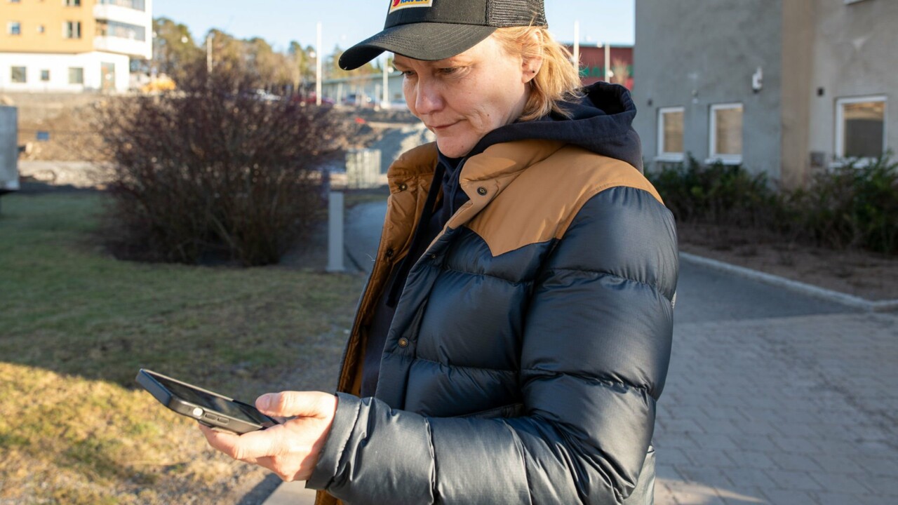 En kvinna står utomhus och tittar på sin mobil som hon håller i handen.