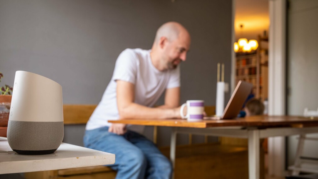 Man som sitter vid sin dator i köket, i förgrunden syns en smart högtalare.