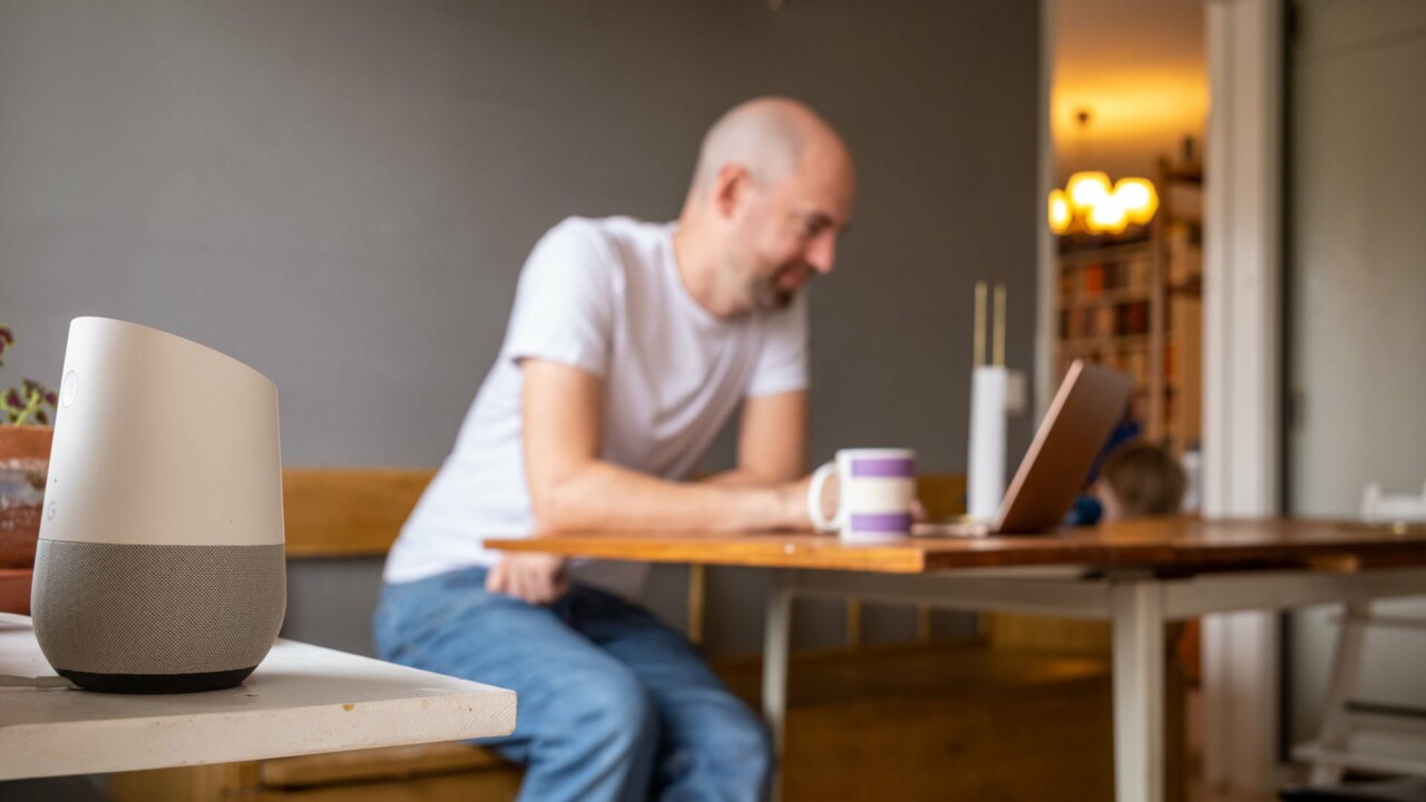 Man som sitter vid sin dator i köket, i förgrunden syns en smart högtalare.