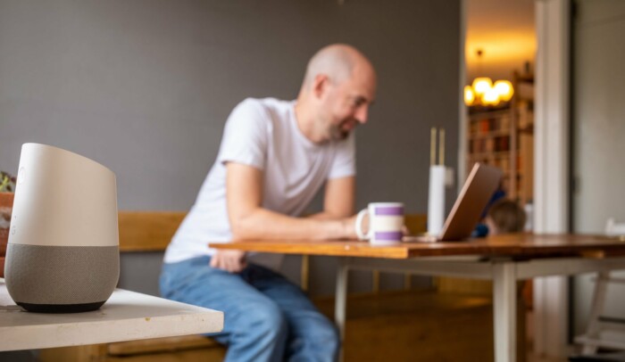 Man som sitter vid sin dator i köket, i förgrunden syns en smart högtalare.