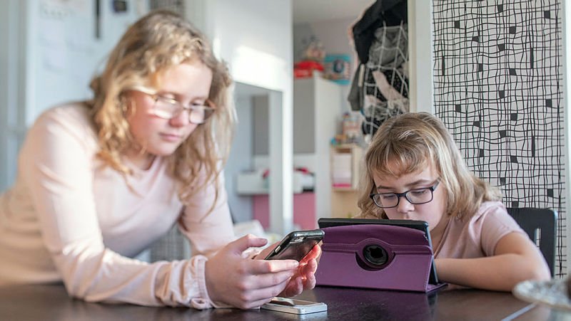 Två flickor sitter vid ett matbord med varsin skärm.