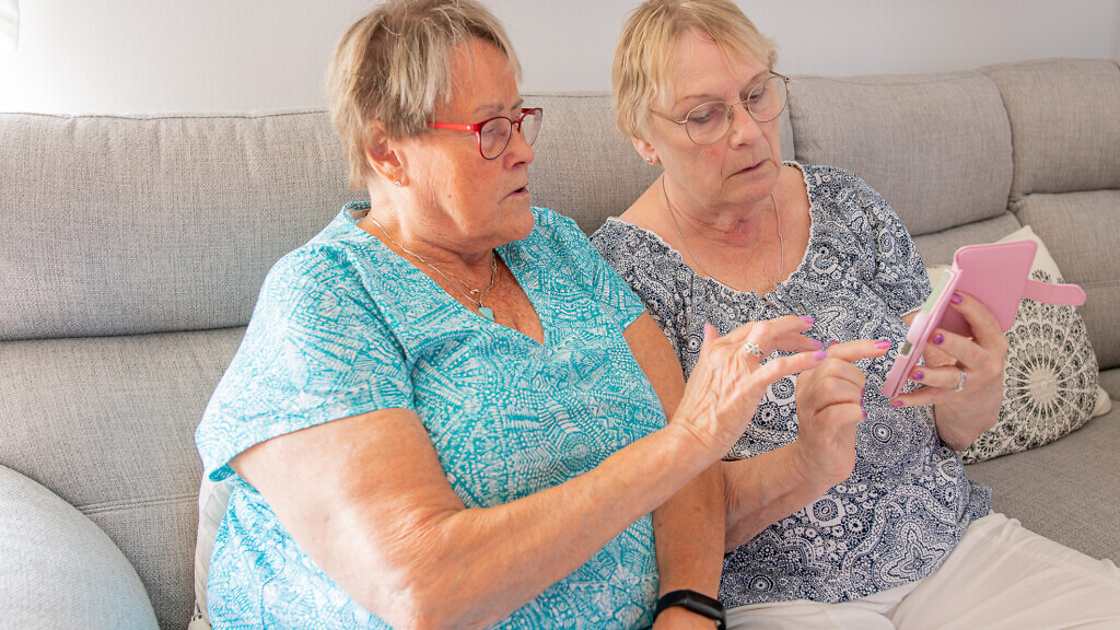 Två kvinnor sitter i en soffa och tittar på en mobil. Idag sker många bedrägerier på internet.
