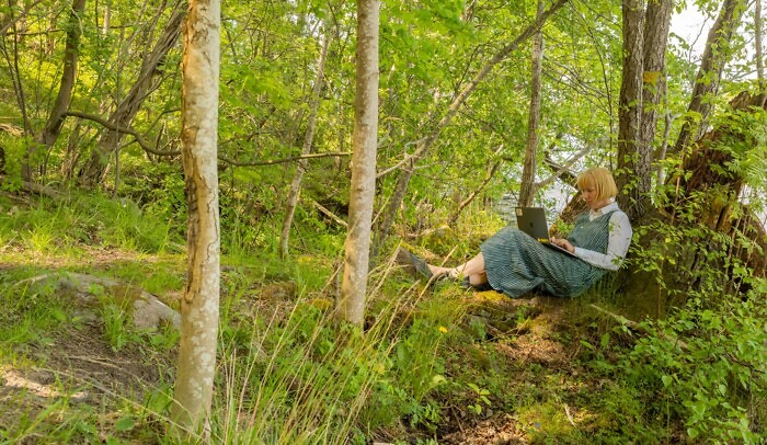 Kvinna sitter ute i naturen med en laptop i knät.