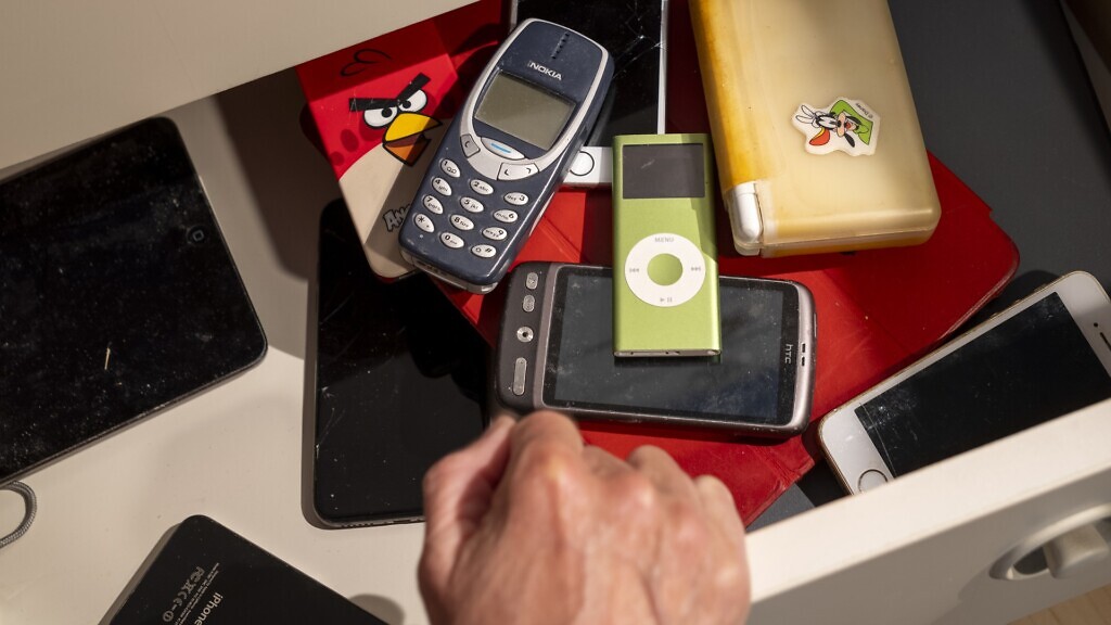 En hand öppnar en låda, i lådan finns massor av gamla digitala enheter så som mobiler och ipods.