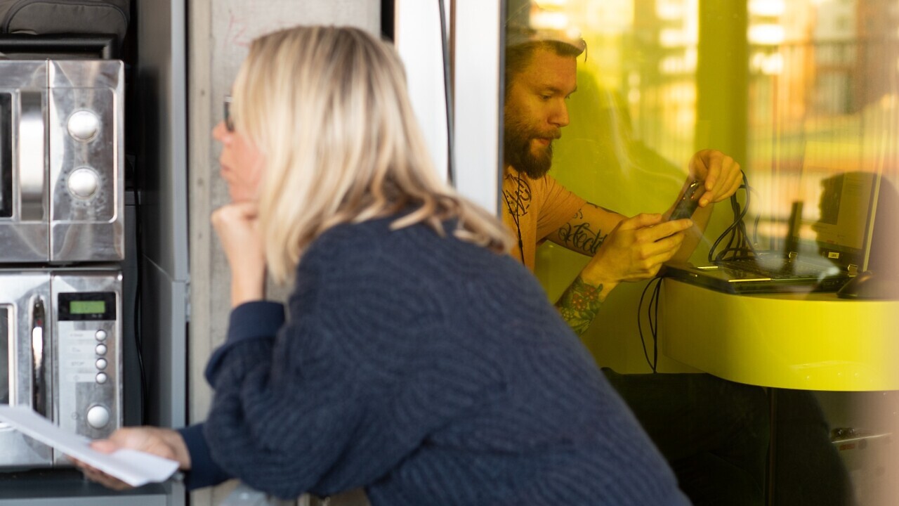 Person med mobiltelefon, dator och hörlurar sitter bakom gul glasvägg på Goto10. Framför skärmen sitter en kvinna i publiken till ett event om övervakningsteknik. Mikrovågsugnar syns bakom henne till vänster i bilden.