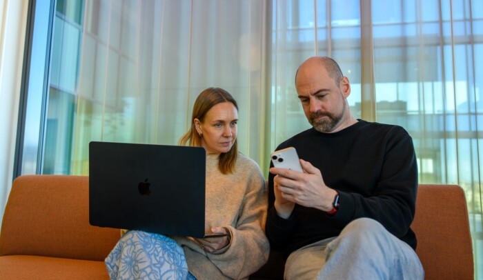 Två personer sitter i en soffa och kollar på en dator och en mobiltelefon.