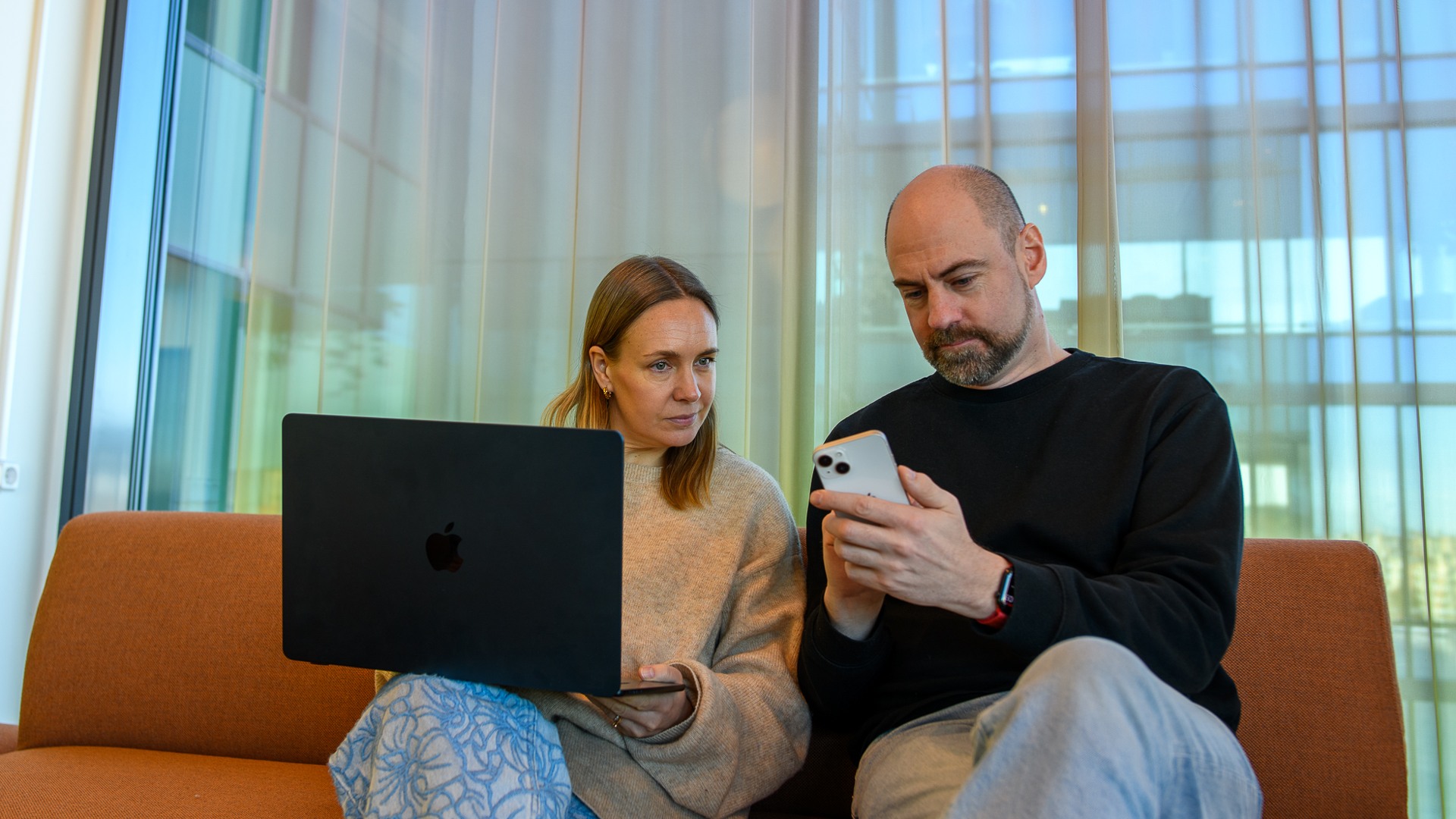 Två personer sitter i en soffa och kollar på en dator och en mobiltelefon.