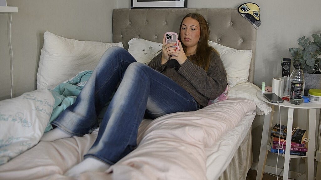 Ung flicka sitter på sin säng och kollar mobiltelefonen.
